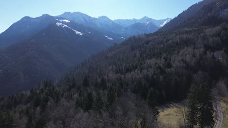 Drone-fly-along-mountain-road-above-pine-forest-with-snow-covered-summit-in-background