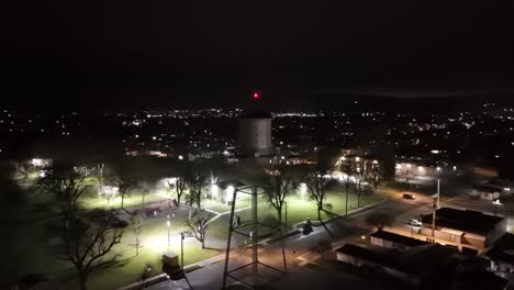 Drone-flies-orbit-around-small-town-water-tank-next-to-green-city-park-at-night