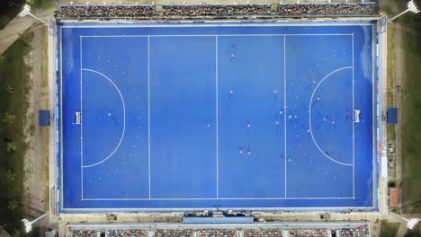 Toma-Aérea-Cenital-De-Un-Campo-De-Hockey-Sobre-Césped-Azul-Y-Puestos-Con-Gente-Donde-Se-Juega-Un-Partido.