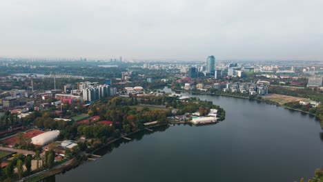 Floreasca-lake-with-bucharest-skyline,-serene-waters,-urban-landscape,-cloudy-day,-cityscape-backdrop,-aerial-view