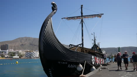 Réplica-Turística-De-Un-Barco-Vikingo-Atracado-En-Un-Puerto-Moderno,-Turistas-Caminando,-Con-Paisaje-Urbano-Y-Montañas-Al-Fondo