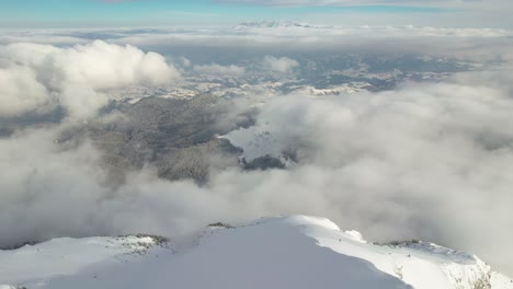 Aerial-view-over-Piatra-Craiului-Mountains,-snow-covered-peaks-piercing-through-white-clouds