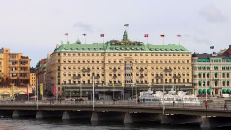 Grand-Hotel-facade-in-Stockholm-with-flags-waving,-cars-on-bridge,-cloudy-sky