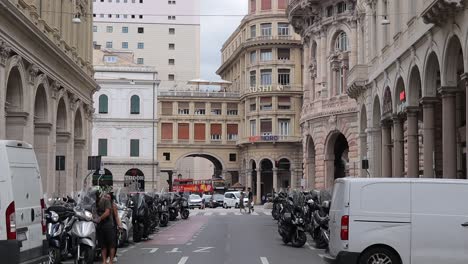 Slow-motion-clip-panning-down-showing-busy-city-street-in-Genoa,-Italy,-with-traffic,-parked-vehicles-and-pedestrians-crossing-the-street