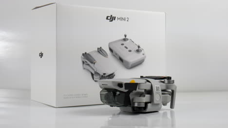 Nuevo-Drone-Dji-Mini-2-Plegado-Junto-A-La-Caja-Del-Paquete,-Foto-De-Estudio-Con-Fondo-Blanco