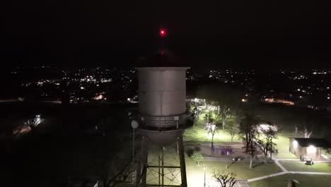 Drone-flies-very-close-to-a-small-town-water-tank-next-to-a-green-grass-city-park-at-night