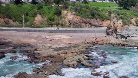 Gente-Y-Un-Pescador-En-Las-Rocas-Mientras-Las-Olas-Llegan-Y-La-Gente-Camina-Por-El-Sendero-Más-Allá.