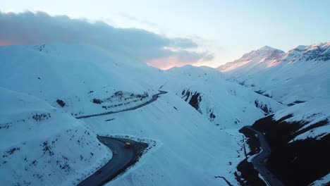 Disparo-Aéreo-De-Drones-Sobrevolando-Una-Montaña-Cubierta-De-Nieve-Mirando-El-Maravilloso-Paisaje-Rojo-Del-Atardecer-Y-Un-Camino-Sinuoso-En-El-Campo-Viajes-De-Invierno-En-La-Temporada-De-Nieve-Fría-En-La-Naturaleza-Salvaje-El-Paisaje-Escénico