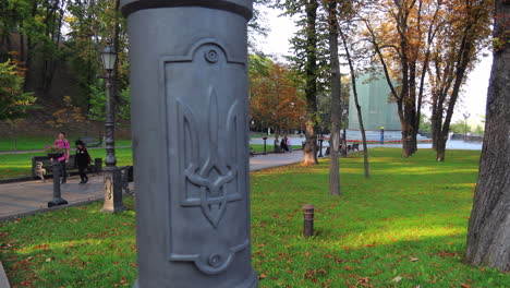 Park-with-traditional-Coat-of-arms-of-Ukraine-near-the-Glass-Bridge-in-Kyiv-city-center-Ukraine,-people-enjoying-the-nature,-green-grass-and-trees,-4K-static-shot