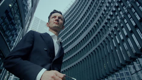 Elegant-Handsome-Business-Man-In-Black-Suit-Standing-Outdoors-Taking-Off-His-Glasses,-Cinematic-Low-Angle
