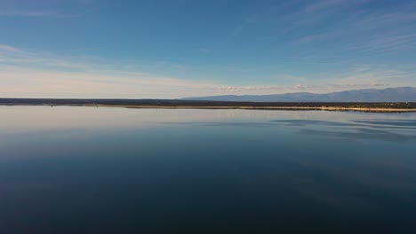 Seitlicher-Flug-Mit-Einer-Drohne-In-Einem-Stausee-An-Einem-Winternachmittag-Mit-Bergen-Im-Hintergrund-Sehen-Wir-Die-Reflexion-Des-Blau-weißen-Himmels-Im-Wasser,-Als-Wäre-Es-Ein-Spiegel,-Alles-Ist-Ruhig