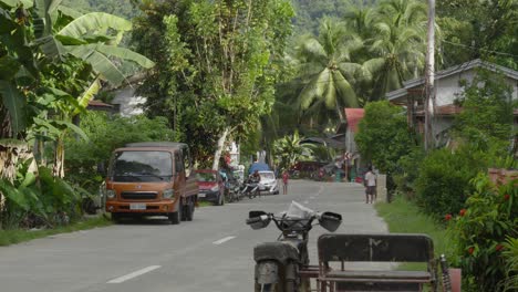 a-tropical-jungle-asphalted-street-in-Philippines,-people-walk-by-old-cars-in-full-greenery,-banana-trees-and-tropical-cinematic-village-feeling