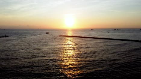 Golden-sunrise-over-calm-sea-with-silhouettes-of-distant-ships-and-breakwater,-aerial-view