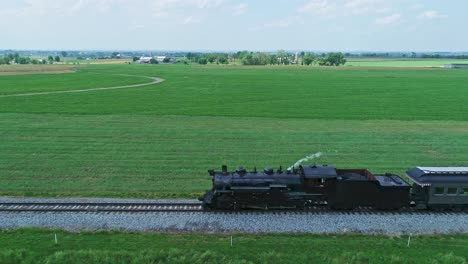 An-Aerial-Side-View-of-a-Steam-Passenger-Train,-Blowing-Smoke-and-Steam-Traveling-on-a-Single-Track-in-Rural-Country,-on-a-Partially-Sunny-Day