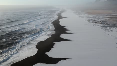 Cinematic-drone-shot-showing-foam-of-ocean-covering-black-basalt-beach-on-Iceland