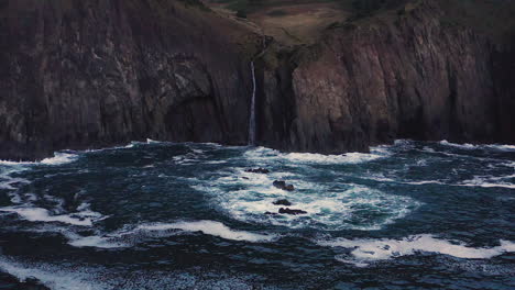 Flyover-past-Oregon-Coast-waterfall-cascading-into-turbulent-Pacific-Ocean