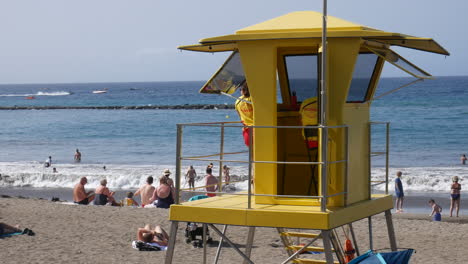 Sonnenanbeter-Und-Schwimmer-Genießen-Das-Meer-In-Der-Nähe-Einer-Auffälligen-Gelben-Rettungsschwimmerstation-Mit-Rollenden-Wellen-Und-Entspannter-Strandatmosphäre