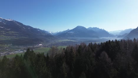 Aerial-view-over-Nenzing-Austria,-Drone-fly-above-pine-forest-revealing-cityscape-with-snow-capped-mountains-in-background-on-a-beautiful-sunny-day-with-clear-blue-sky