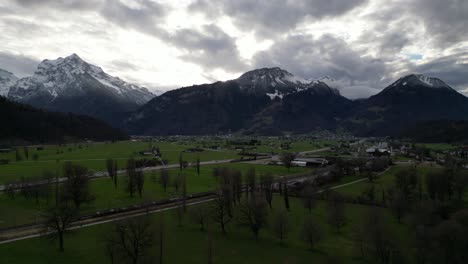 Aerial-pan-across-lush-green-farm-fields-and-leafless-trees-below-swiss-mountains-in-Walensee