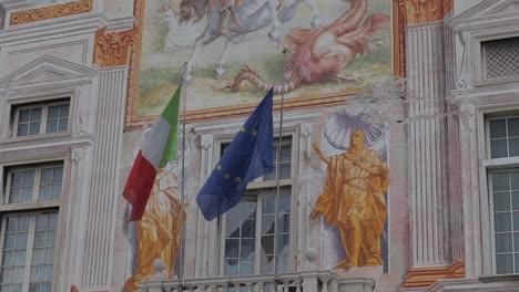 Primer-Plano-De-Murales-Detallados-En-El-Palacio-San-Giorgio,-Génova,-Italia,-Que-También-Muestran-Banderas-Italianas-Y-De-La-UE-Ondeando-En-La-Brisa.