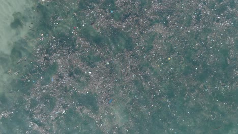 Drone-Descendente-De-Arriba-Hacia-Abajo-A-Gran-Altitud-De-Agua-Contaminada-Llena-De-Basura-Flotando-Sobre-Arrecifes-De-Coral-Muertos-En-Las-Aguas-Tropicales-Turquesas-De-Bali-Indonesia