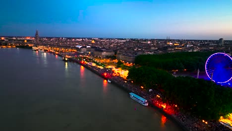 Large-ferris-wheel-lit-up-in-neon-lights-with-crowds-in-Bordeaux-France-during-wine-fair,-Aerial-pan-right-shot