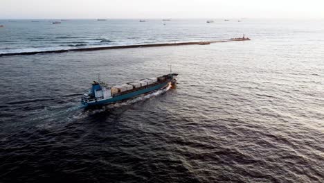 Cargo-ship-cruising-through-ocean-at-sunset,-aerial-view,-with-other-vessels-on-horizon
