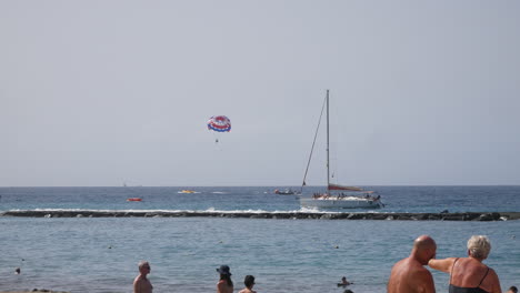 Beach-visitors-enjoy-the-sun-as-a-parasailing-parasailer-flies-over-the-sea-near-a-sailboat,-with-a-scenic-breakwater-under-the-clear-blue-sky
