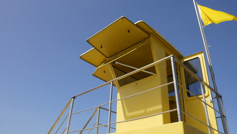 Una-Torre-De-Salvavidas-Amarilla-Contra-Un-Cielo-Azul-Claro,-Una-Bandera-Ondeando-Con-La-Brisa,-Un-Símbolo-De-Seguridad-Y-Vigilancia-En-Verano-En-Las-Playas-Soleadas