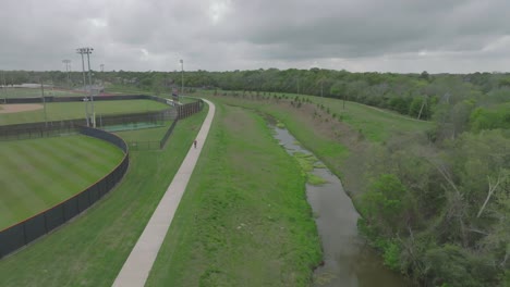 Una-Vista-De-Drone-De-Un-Hombre-Adulto-Andando-En-Bicicleta-Por-El-Sendero-Para-Caminatas-Y-Bicicletas-En-El-área-De-La-Bahía-Detrás-De-La-Escuela-Secundaria-De-Clear-Lake-En-Un-Día-Nublado-En-Clear-Lake,-Houston,-Texas