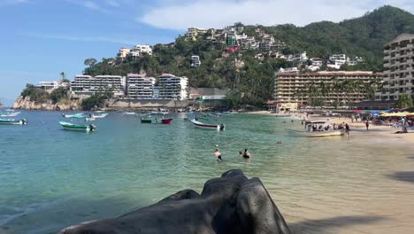 People-swim-in-clean-sea-water-of-Mismaloya-Mexican-beach-hotels-in-green-hill-Hand-Held-shot-of-boats,-blue-ocean-and-skyline,-Mexico-travel-destination