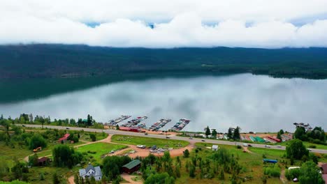 Cars-Driving-by-Breathtaking-Mountain-Lake-Shoreline-with-Rolling-Coulds-in-the-Background-and-Glass-Calm-Reflective-Water