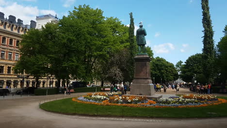 Esplanadi-Park-in-Downtown-Helsinki,-Finland,-Buildings,-Statue-and-People-Walking-on-Spring-Day
