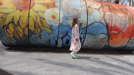 Fixed-shot-of-lady-wearing-casual-floral-clothing,-walking-left-to-right-in-front-of-colourful-urban-mural