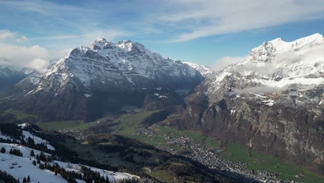 Aerial-bird's-eye-view-above-village-at-base-of-green-valley-with-snow-and-cloud-covered-mountains