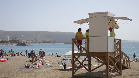 Una-Animada-Escena-De-Playa-Con-Un-Salvavidas-De-Guardia,-Supervisando-La-Seguridad-De-Los-Bañistas-Y-Bañistas-Frente-A-Un-Pintoresco-Telón-De-Fondo-Montañoso-En-Los-Christianos-En-Tenerife.