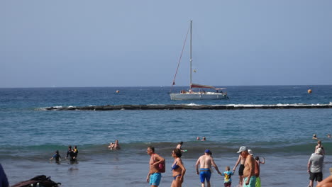 Strandbesucher-Schwimmen-Und-Spazieren-Am-Wasser-Entlang,-Während-Ein-Segelboot-Unter-Freiem-Himmel-An-Einem-Felsigen-Wellenbrecher-Vorbeifährt-Und-So-Den-Küstenurlaub-Einfängt