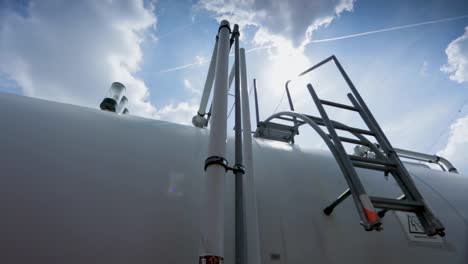 Low-angle-view-of-a-white-industrial-tank-with-ladders-against-a-blue-sky,-sun-flare-effect