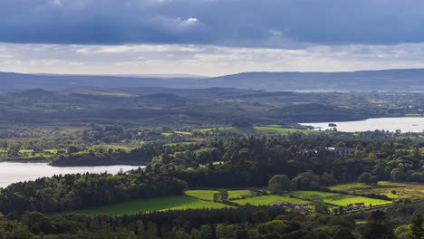 Time-lapse-of-rural-landscape-with-Kilronan-castle-in-the-distance-surrounded-by-lake,-forest-and-hills-during-a-cloudy-day-viewed-from-above-Lough-Meelagh-in-county-Roscommon-in-Ireland