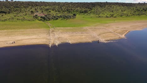 Vuelo-Hasta-El-Borde-De-Un-Embalse-Donde-Vemos-Los-Restos-De-Piedra-De-Unas-Estructuras-Antiguas-De-Algo-Que-Ya-Ha-Desaparecido,-Un-Antiguo-Camino-Inundado-Por-El-Embalse,-Terminamos-En-Tierra-Verde