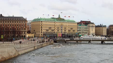 Blick-Auf-Das-Wasser-Des-Grand-Hotels-In-Stockholm-Mit-Wehenden-Fahnen,-Bewölktem-Himmel-Darüber-Und-Fußgängern