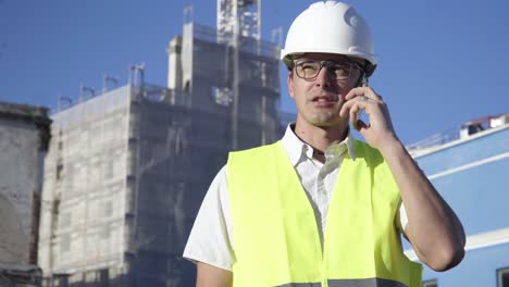 At-the-construction-site,-a-young-engineer-wearing-glasses-and-a-white-helmet-is-having-a-conversation-on-the-phone