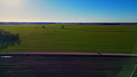 Luftaufnahme-Einer-Minimalistischen-Landschaft-Bei-Sonnenuntergang-Mit-Einem-Baum-Auf-Einem-Grünen-Landwirtschaftlichen-Feld-Und-Einem-Auto,-Das-Auf-Einer-Nebenstraße-Auf-Dem-Land-Fährt