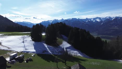Drone-fly-above-field-of-snow-revealing-green-grass-hills-and-farm-yard-on-the-mountains-with-amazing-view-over-snowcapped-mountains-on-a-sunny-day