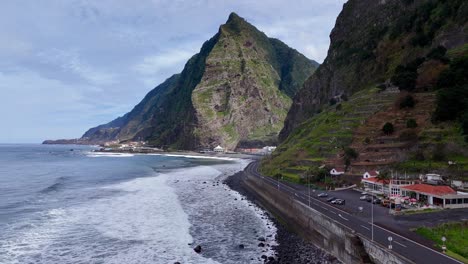 Drone-footage-near-the-Ribeiro-do-Inferno-in-Madeira-island-on-a-sunny-day-with-waves-hitting-the-road-and-a-mountain-in-the-background