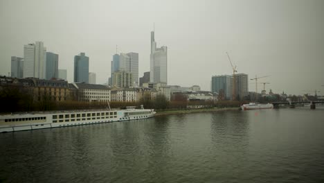 Overcast-view-of-Frankfurt-skyline-with-Main-River-and-passing-ships,-cranes-at-construction-sites