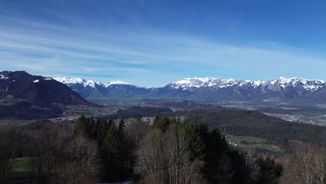 Drone-fly-above-forest-and-revealing-amazing-cityscape-with-winter-mountain-landscape-and-snowcapped-mountains-on-a-sunny-day-in-Feldkirch,-Austria