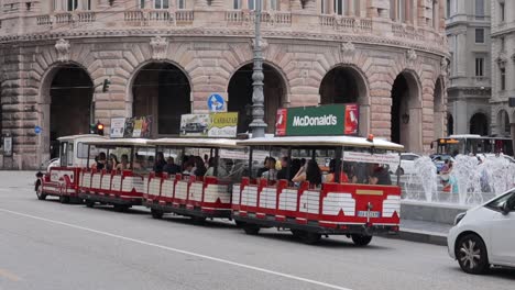 Red-and-white-tourist-train-driving-through-traditional-Italian-town,-with-advertising-boards-on-top,-full-of-tourists