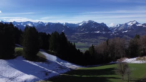 Drone-fly-towards-forest-with-town-and-winter-mountain-landscape-with-snowcapped-mountains-in-background-on-a-sunny-day-sky-in-Austria