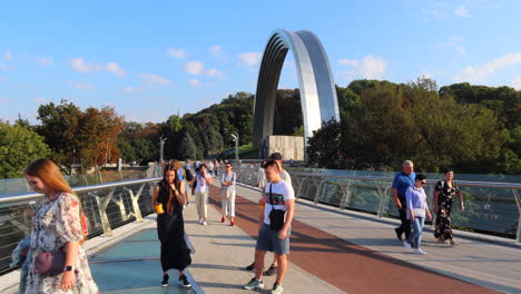 People-walking-on-the-famous-Glass-Bridge-in-Kyiv-city-center-Ukraine,-The-Arch-of-Freedom-of-the-Ukrainian-People,-sunny-weather-and-blue-sky,-bridge-with-glass-floor-panels,-4K-shot